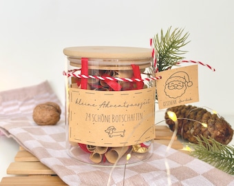 Op de afbeelding: Een doorzichtige glazen pot met een houten deksel gevuld met opgerolde papieren briefjes en rood lint. Het beige etiket bevat de tekst "kleine Adventsauszeit 24 SCHÖNE BOTSCHAFTEN" met een teckel-icoon. Een label toont een illustratie van de Kerstman.