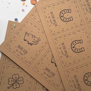 May include: A stack of brown paper cards with black line drawings of a horseshoe, pig, and four-leaf clover. The cards have text in German. Confetti and coins are scattered around the cards.