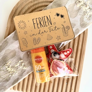 Puede incluir: Una bolsa de plástico transparente que contiene una botella de protector solar amarillo Zenova SPF 50 y una galleta roja con forma de estrella de mar. Una etiqueta de cartón marrón lleva el texto "FERIEN in der Tüte" e iconos veraniegos.