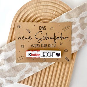 Op de afbeelding: Een bruine kaart met de tekst "DAS neue Schuljahr WIRD FÜR DICH" en een Kinder-chocolade reep met de tekst "Kinder LEICHT" op een houten achtergrond.