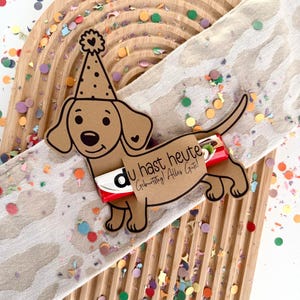 Puede incluir: Un perro salchicha de cartulina marrón con un gorro de fiesta, sosteniendo una barra de caramelo, y el texto "Du hast heute Geburtstag! Alles Gute!" en una bandeja de madera en forma de arcoíris con confeti.