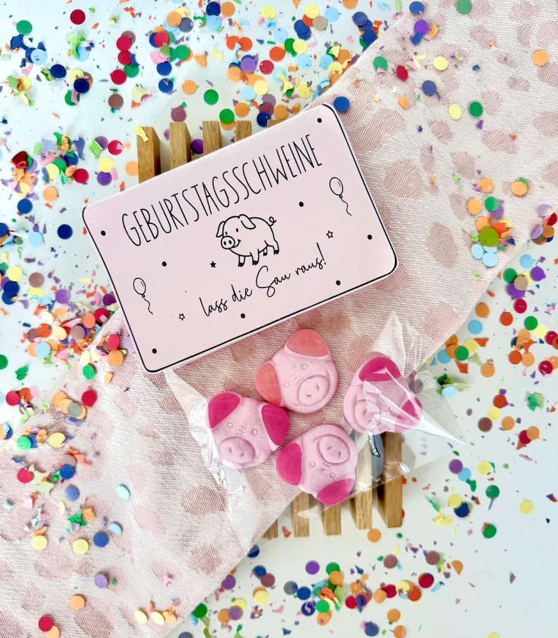 May include: A pink rectangular box with the text "GEBURTSTAGSSCHWEINE" and a pig illustration, accompanied by pink pig-shaped candies in a clear bag. The candies are on a wooden stand, surrounded by colorful confetti.