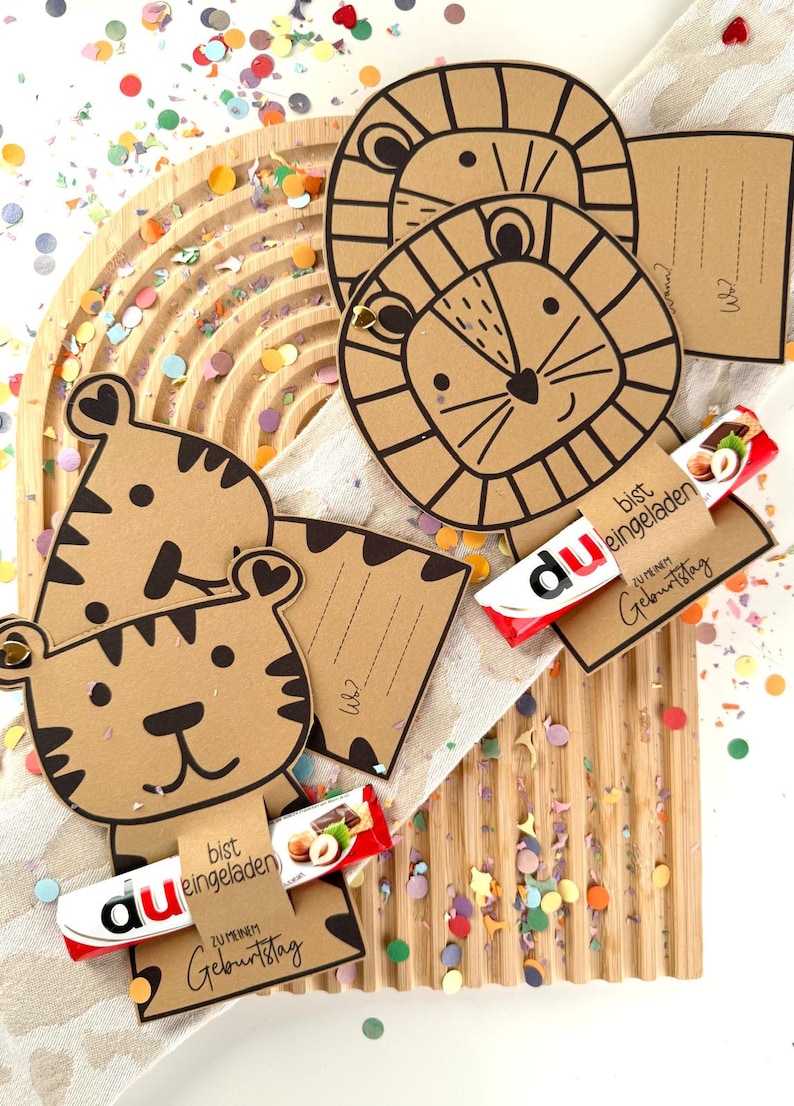 May include: Brown paper animal-shaped birthday cards with black outlines, featuring a tiger and lion design. Each card holds a small chocolate bar with German text. Colorful confetti surrounds the cards on a wooden surface.