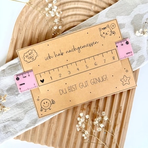 Puede incluir: Una tarjeta rectangular de color canela con el texto en alemán 'ich hab nachgemessen' y 'DU BIST GUT GENUG!', una regla impresa de 1 a 8 cm, una ilustración de un gato y una cara sonriente, sujeta por una cinta métrica rosa de 5 cm.