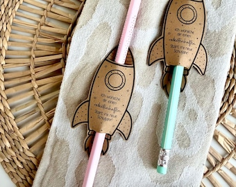 May include: Two pencils, one light pink and one light blue, each inserted through a brown paper rocket-shaped cutout. The rockets feature the German text 'Ich wünsche dir einen raketenhaften Start ins neue Schuljahr' and small star illustrations.