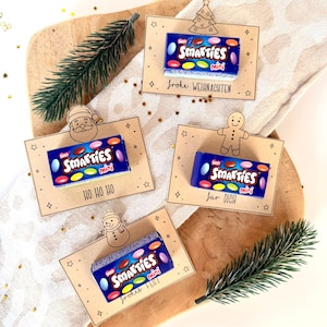 Op de afbeelding: Vier mini Smarties snoepdozen op decoratieve kaarten met vakantie-thema toppers. De kaarten hebben Duitse tekst, waaronder "Frohe Weihnachten", "Ho Ho Ho", "fur DICH" en "frohes Fest". De snoepjes zijn gerangschikt op een houten plank met groen.