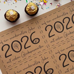 May include: A kraft paper card with the year 2026 printed in black, along with the phrase "auf ein wundervolles Jahr" and "mit vielen Gl&uuml;cksmomenten". Two gold-wrapped chocolate candies and colorful confetti are scattered around the card.