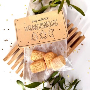 May include: A package of shortbread cookies with intricate designs, presented on a linen napkin with sprigs of green mistletoe. A brown tag reads "aus meiner WEIHNACHTSBÄCKEREI" with festive illustrations. The cookies are in a clear plastic bag.