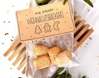 Op de afbeelding: Een pakketje shortbread koekjes met ingewikkelde patronen, gepresenteerd op een linnen servet met groene maretak takjes. Een bruin label leest "aus meiner WEIHNACHTSBÄCKEREI" met feestelijke illustraties. De koekjes zitten in een doorzichtige plastic zak.