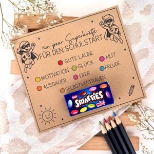 May include: A brown card with the text "ein paar Superkräfte FÜR DEN SCHULSTART" and superhero illustrations. Includes a box of Smarties Mini sweets and coloured pencils. The card lists qualities like "Gute Laune" and "Motivation".