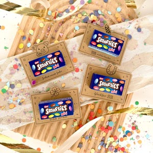 Puede incluir: Cajas de caramelos Mini Smarties sobre tarjetas decorativas con confeti. Cada tarjeta tiene un diseño diferente, incluyendo un cerdo, una herradura y un trébol de cuatro hojas. Las tarjetas tienen texto en alemán, incluyendo "Frohes neues Jahr".