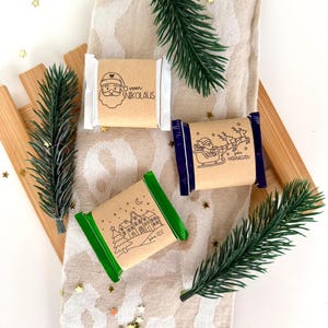 Könnte beinhalten: Drei verpackte Schokoriegel mit festlichen Designs. Einer zeigt den Weihnachtsmann mit dem Text "vom Nikolaus", ein anderer zeigt den Weihnachtsmann im Schlitten mit Rentieren, und der dritte zeigt eine Dorflandschaft. Die Riegel liegen auf einem gemusterten Stoff mit Tannenzweigen.