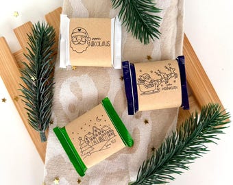 May include: Three wrapped chocolate bars with festive designs. One features Santa Claus with the text "vom Nikolaus", another shows Santa in a sleigh with reindeer, and the third depicts a village scene. The bars are on a patterned cloth with pine sprigs.