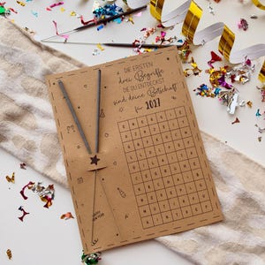May include: A rectangular brown card featuring a word search puzzle with the German text DIE ERSTEN drei Begriffe DIE DU ENTDECKST sind deine Botschaft für 2027. Two thin grey sparklers are attached to the left side with a small star cutout.