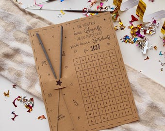 May include: A rectangular brown card featuring a word search puzzle with the German text DIE ERSTEN drei Begriffe DIE DU ENTDECKST sind deine Botschaft für 2027. Two thin grey sparklers are attached to the left side with a small star cutout.