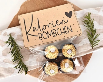 Può includere: Un piccolo sacchetto di carta marrone con un cuore e il testo "Kalorien-BÖMBCHEN" su di esso. Il sacchetto contiene quattro caramelle di cioccolato avvolte in oro.