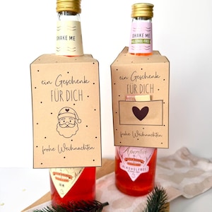 Puede incluir: Dos pequeñas botellas de vidrio llenas de líquido rojo, cada una con una tapa dorada y una etiqueta decorativa. Las etiquetas dicen "Shake Me" y "ein Geschenk FÜR DICH" con una ilustración de Papá Noel y "frohe Weihnachten".