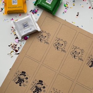 May include: An overhead shot features three Ritter Sport chocolate bars in yellow, green, and white packaging, alongside a sheet of brown paper with printed illustrations. The paper has images of pigs, chimney sweeps, and champagne glasses. Confetti is scattered around the scene.