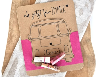 May include: A brown card with a black and white illustration of a vintage bus. The card says "ab jetzt für IMMER" with a heart symbol. The card is wrapped in a pink ribbon and has rolled up bills tied with rubber bands.