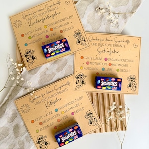 Puede incluir: Tres tarjetas rectangulares de color tostado con texto negro e ilustraciones de superhéroes. Cada tarjeta incluye la frase Danke für deine Superkräfte y una pequeña caja de caramelos Smarties Mini. El texto enumera rasgos positivos.