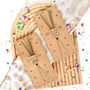 以下が含まれることがあります： スパークラーが取り付けられた茶色の厚紙タグ2枚。タグのテキストは「Bring das neue Jahr zum Leuchten」と「Frohes neues Jahr」です。