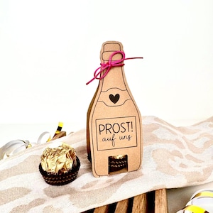 Könnte beinhalten: Eine braune Papp-Champagnerflaschen-Geschenkbox mit einem rosa Band und dem Text "PROST! auf uns" auf der Vorderseite. Die Box ist so konzipiert, dass sie eine kleine Schokolade aufnehmen kann.