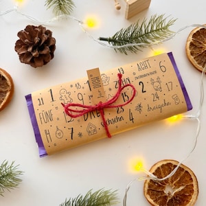 Könnte beinhalten: Eine rechteckige Schokoladentafel in braunem Papier mit schwarzen Zahlen und weihnachtlichen Illustrationen, gebunden mit roter Kordel und einem Anhänger mit der Aufschrift "für DICH". Das Papier enthält Texte wie "FÜNF", "ACHT", "ZWEIHUNDZWANZIG", "dreizehn" und "sechzehn".