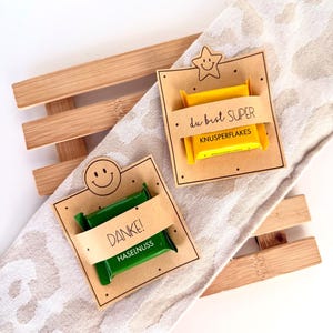 Può includere: Due piccole barrette di cioccolato rettangolari in involucri gialli e verdi, ciascuna fissata su un cartoncino quadrato in carta kraft. Il cartoncino giallo presenta una stella e la scritta "du bist SUPER KNUSPERFLAKES", quello verde uno smiley e "DANKE! HASELNUSS."