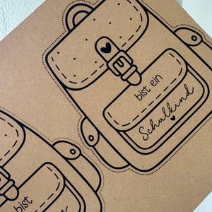 May include: Brown paper with a black line drawing of a backpack. The backpack design includes a buckle, heart, and the words "bist ein Schulkind" in script. The design is repeated on the paper.