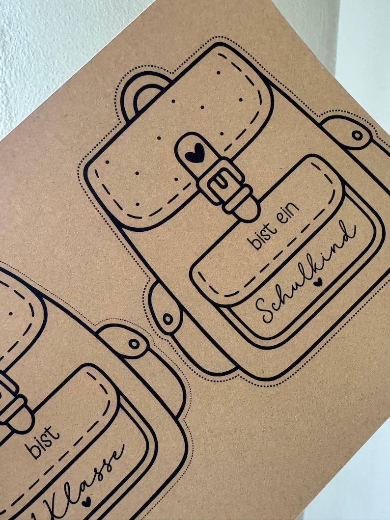 May include: Brown paper with a black line drawing of a backpack. The backpack design includes a buckle, heart, and the words "bist ein Schulkind" in script. The design is repeated on the paper.