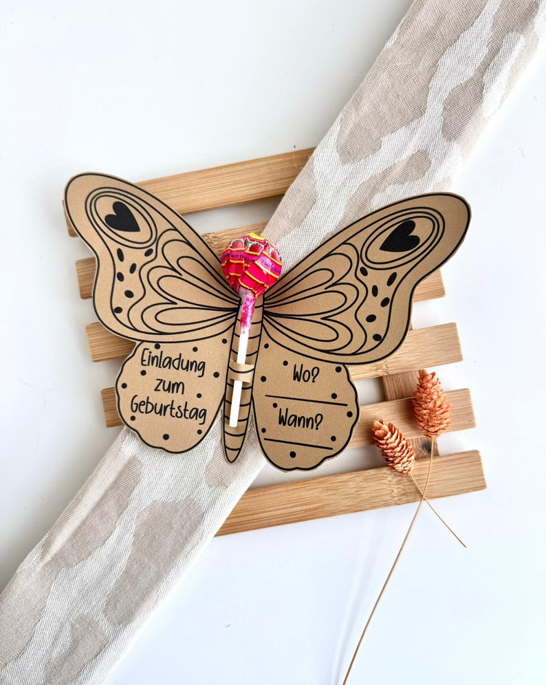 K&ouml;nnte beinhalten: Ein brauner Papier-Schmetterling mit schwarzen Umrissen und Herzen. Der Schmetterling hat einen Lolli daran befestigt. Der Schmetterling hat den Text "Einladung zum Geburtstag" auf einem Fl&uuml;gel und "Wo? Wann?" auf dem anderen Fl&uuml;gel.