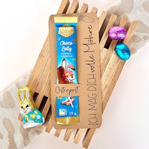 May include: A brown wooden Easter basket containing a chocolate bar, a bunny-shaped chocolate, and two foil-wrapped chocolate eggs. The chocolate bar is wrapped in blue foil with a cartoon bunny and the text "Oster Phantasie Choco Lolly Vollmilch-Schokolade". The basket has a brown paper tag with the text "ICH MAG DICH volle M&ouml;hre" and "Osterpost".