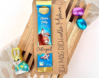 May include: A brown wooden Easter basket with a chocolate bar, a bunny-shaped chocolate, and two foil-wrapped chocolate eggs. The chocolate bar is wrapped in blue foil with a cartoon bunny and the text "Oster Phantasie Choco Lolly Vollmilch-Schokolade". The basket has a brown paper tag with the text "ICH MAG DICH volle Möhre" and "Osterpost".