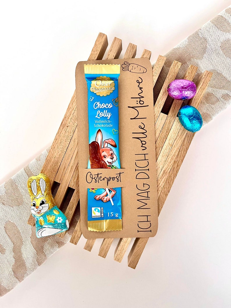 May include: A brown wooden Easter basket containing a chocolate bar, a bunny-shaped chocolate, and two foil-wrapped chocolate eggs. The chocolate bar is wrapped in blue foil with a cartoon bunny and the text "Oster Phantasie Choco Lolly Vollmilch-Schokolade". The basket has a brown paper tag with the text "ICH MAG DICH volle M&ouml;hre" and "Osterpost".
