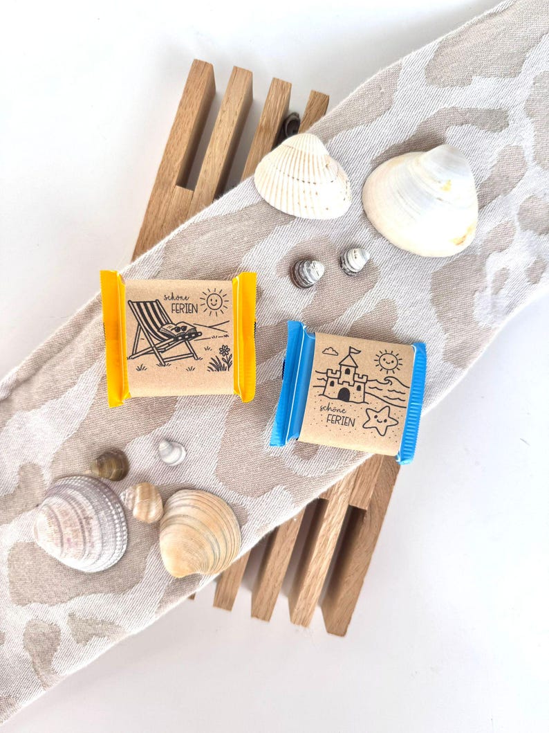 May include: Two small wrapped chocolate bars with beach-themed illustrations and the text "schone FERIEN" are displayed with seashells on a patterned cloth. One bar has a yellow wrapper and the other has a blue wrapper. A wooden rack is also in the image.