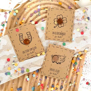 May include: Three small, brown cards with lucky symbols: a horseshoe, a four-leaf clover, and a pig. Each card has a penny attached and is decorated with black dotted lines and text in German. Confetti and ribbons surround the cards.
