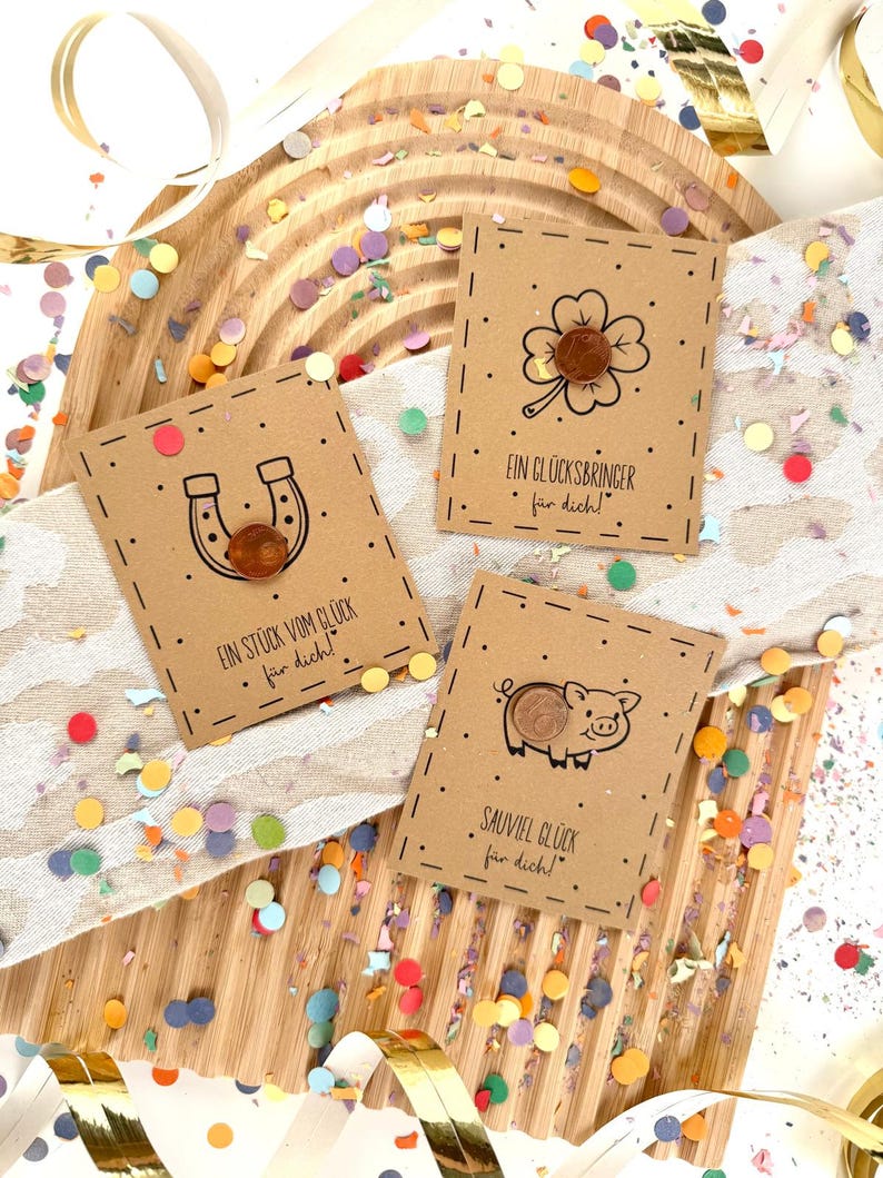 May include: Three small, brown cards with lucky symbols: a horseshoe, a four-leaf clover, and a pig. Each card has a penny attached and is decorated with black dotted lines and text in German. Confetti and ribbons surround the cards.