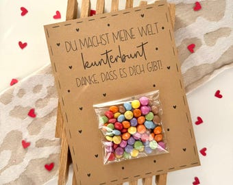 Op de afbeelding: Een bruine kaart met de tekst "DU MACHST MEINE WELT. kunterbunt. DANKE, DASS ES DICH GIBT!" omringd door kleine rode hartjes. Een klein zakje met kleurrijke snoepjes ligt op de kaart.