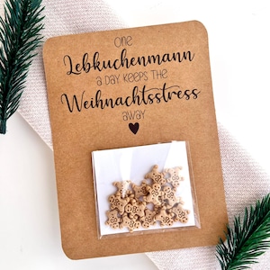 Pu&ograve; includere: Un biglietto di carta kraft con il testo "one Lebkuchenmann a day keeps the Weihnachtsstress away" e un piccolo sacchetto di decorazioni a forma di omino di pan di zenzero. Il biglietto &egrave; decorato con rametti di sempreverde.