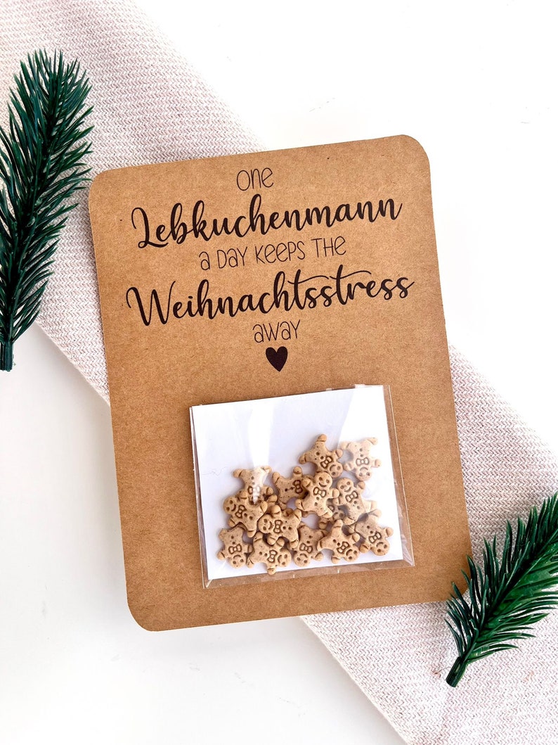 Pu&ograve; includere: Un biglietto di carta kraft con il testo "one Lebkuchenmann a day keeps the Weihnachtsstress away" e un piccolo sacchetto di decorazioni a forma di omino di pan di zenzero. Il biglietto &egrave; decorato con rametti di sempreverde.