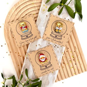 May include: Three Christmas cards with snow globe designs and text. Each card features a different character inside the globe: a teddy bear and Santa Claus. The cards are on a wooden tray with mistletoe.