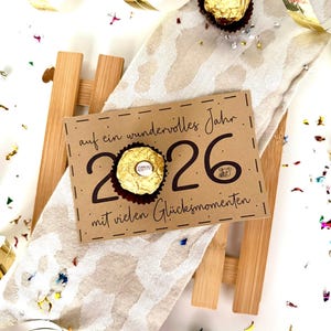以下が含まれることがあります： 2026年のための、お祝いのグリーティングカード。テキストは「auf ein wundervolles Jahr mit vielen Glücksmomenten」と書かれており、2つのチョコレート菓子が添えられています。カードは模様入りの布の上に置かれ、紙吹雪と金色のリボンが飾られています。