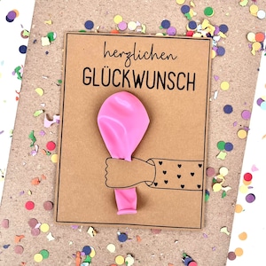 May include: A greeting card with the German text "herzlichen GL&Uuml;CKWUNSCH" and a pink balloon held by a cartoon arm with heart details. The card is on a brown background with colorful confetti.