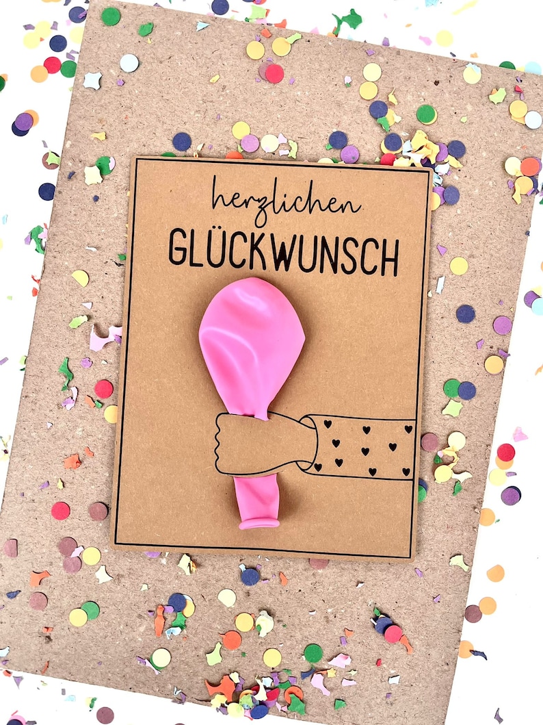 May include: A greeting card with the German text "herzlichen GL&Uuml;CKWUNSCH" and a pink balloon held by a cartoon arm with heart details. The card is on a brown background with colorful confetti.
