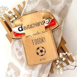 Pode incluir: Um recorte de papel bege em forma de t-shirt com uma barra de chocolate inserida na gola. O texto "deutschland vor, noch ein TOOOR!" está impresso em tinta preta, juntamente com um gráfico de bola de futebol e "WM 2026" na frente.