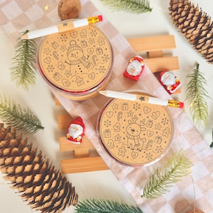 Può includere: Due contenitori rotondi con coperchi color cuoio illustrati con un pupazzo di neve e una renna, ciascuno sormontato da una matita bianca. Sono circondati da pigne, piccole statuette di Babbo Natale e rametti di pino su un tessuto a quadri.