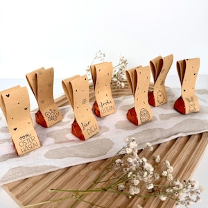 Könnte beinhalten: Osterschokoladen, verpackt in braunem Papier mit verschiedenen Designs, darunter Herzen, Schmetterlinge und Ostereier. Das Papier trägt auch Sätze wie "Frohe Ostern" und "Für dich". Die Pralinen sind auf einem Holzbrett angeordnet.