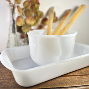 May include: A white ceramic serving set with a rectangular tray and a round bowl. Three wooden sticks are placed inside the bowl. The set is on a wooden surface, with a vase of dried flowers in the background.