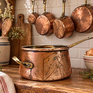May include: A large, round copper pot with a long brass handle and a smaller side handle. The pot has a weathered, metallic finish with visible signs of use and oxidation. It sits on a wooden surface, showcasing a classic, functional kitchen design.