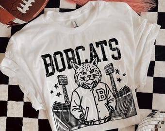 Pode incluir: Camiseta branca com a palavra "BOBCATS" em letras pretas e um lince-vermelho de desenho animado com uma jaqueta. O lince-vermelho está em frente a um estádio. Uma bola de futebol americana marrom e um par de tênis brancos também são visíveis.